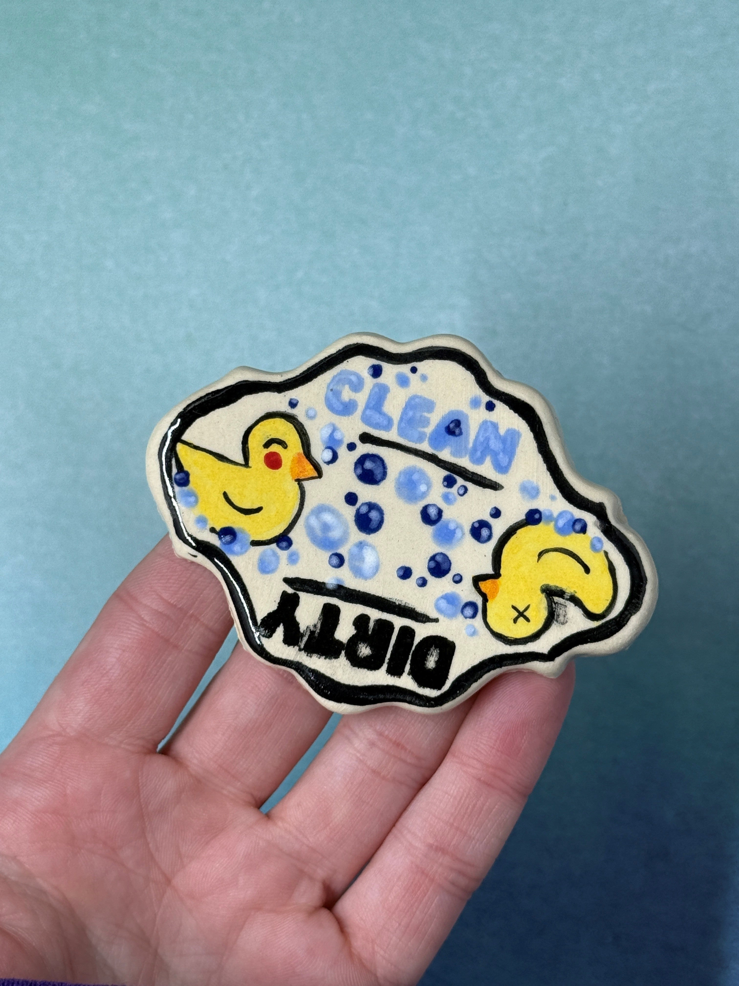 A hand holding a hand-painted ceramic pin featuring two yellow rubber ducks in a soapy bubble outline with the text 'CLEAN DIRTY' on a blue gradient background.